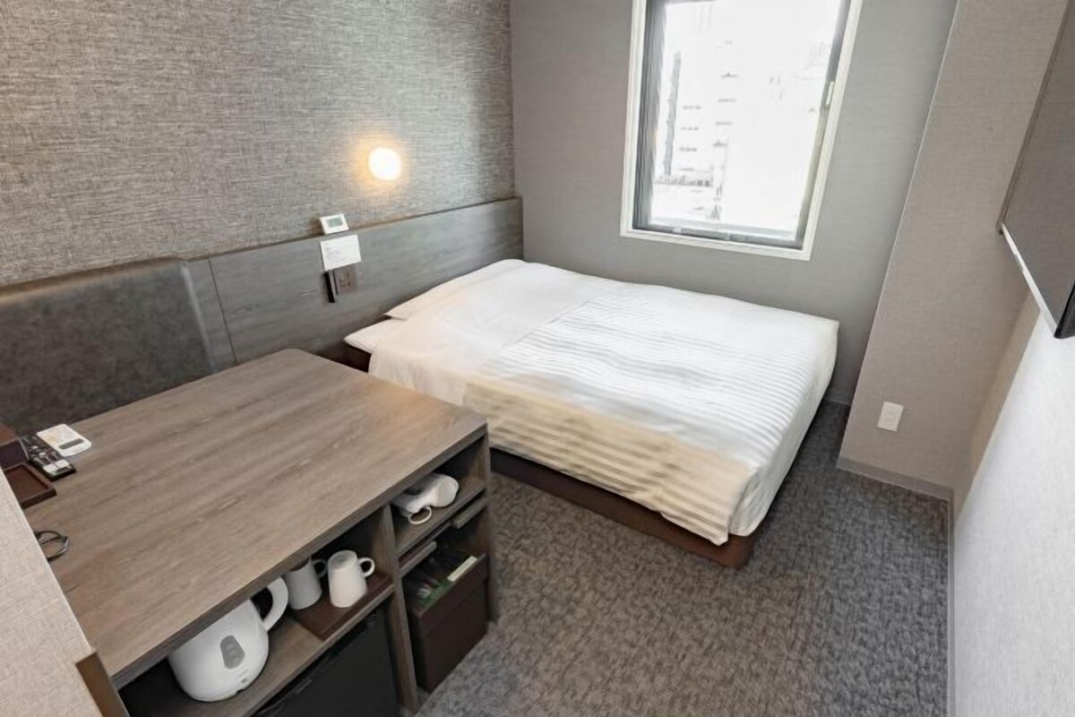A compact hotel room with a bed, a desk with a chair, a shelf with items, a window, and a wall-mounted light.