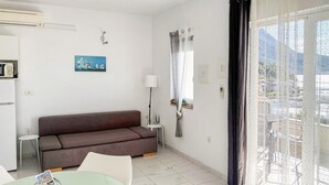 Ein modernes Wohnzimmer mit einem braunen Sofa, einem Glastisch mit Zeitschriften, einer Lampe und Blick auf eine gebirgige Landschaft durch ein Fenster mit transparenten Vorhängen.