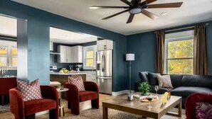 Una sala moderna con una pared de acento azul oscuro, un sofá de cuero negro, sillones de terciopelo rojo, una mesa de centro de madera y una cocina al fondo.