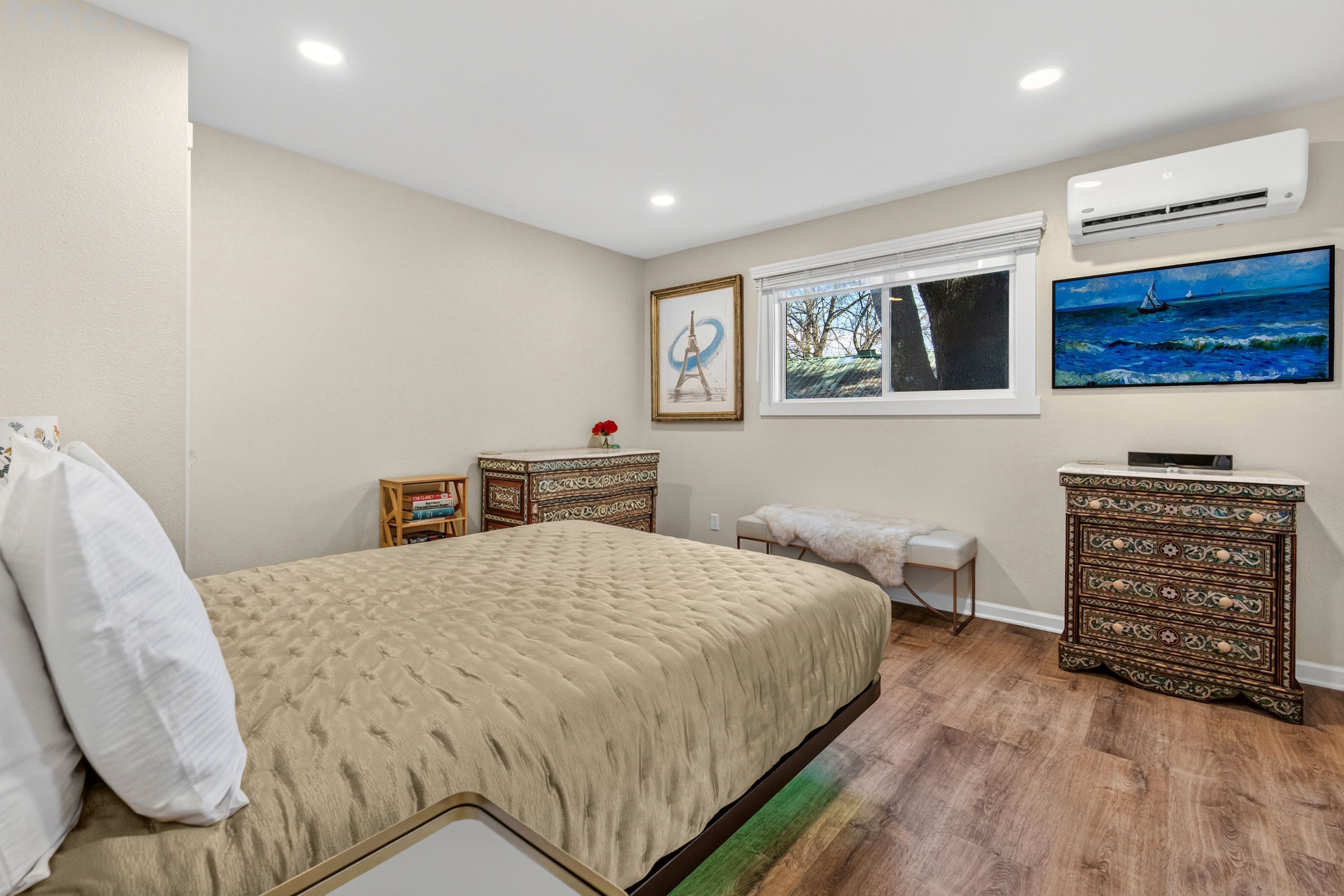 A bedroom with a bed, a dresser, a bench, a window, and artwork on the walls.