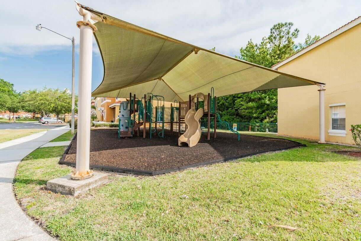 Un terrain de jeux avec un toboggan et des structures d’escalade, protégé du soleil par un auvent, situé devant un bâtiment.