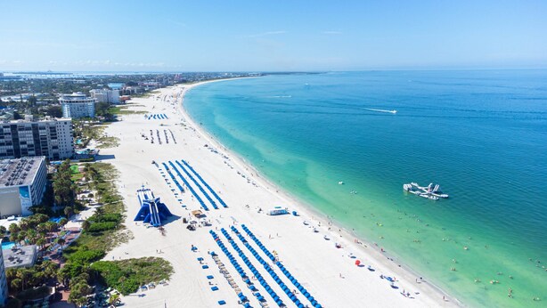 Ein Strand mit Reihen blauer Sonnenschirme und Liegestühle, ein klares blaues Meer und Gebäude entlang der Küste.