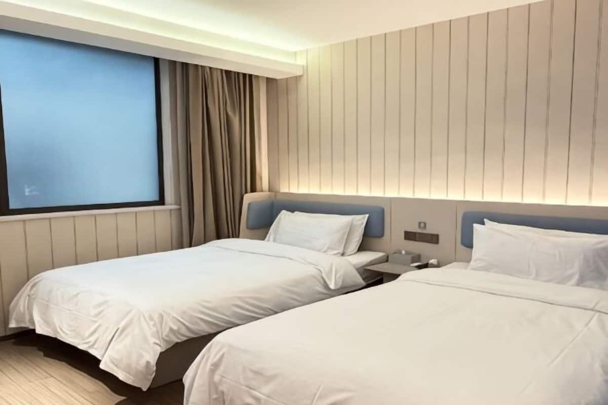 A hotel room with two beds, a window with curtains, a wall-mounted light, and a bedside table.
