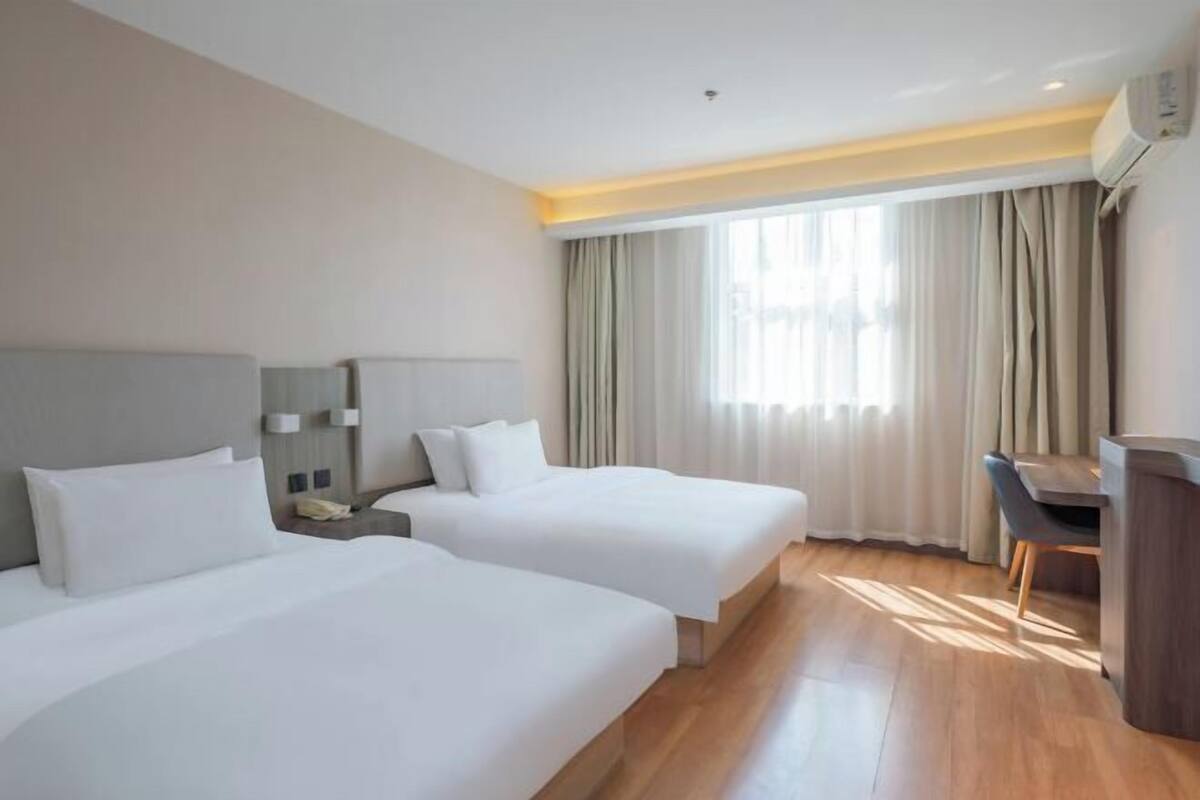 A hotel room with two beds, a desk, a chair, a nightstand, and a window with curtains.