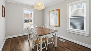 A dining room with a wooden table and white chairs, a hanging light fixture, and a window with blinds.