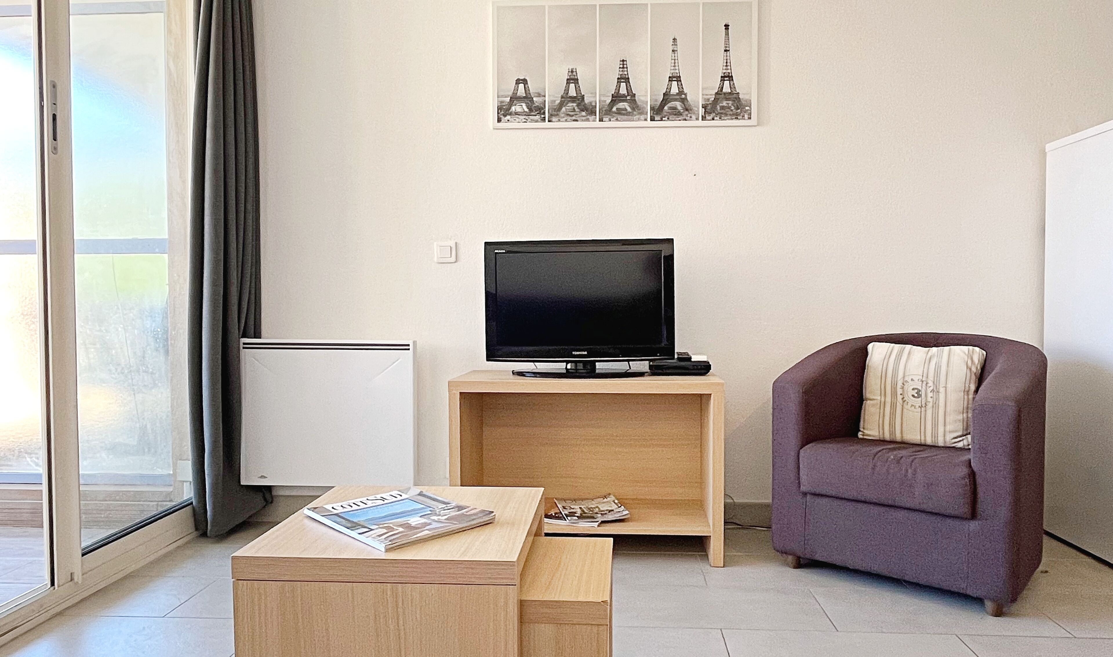 Una sala de estar con un televisor sobre un mueble de madera, un sillón morado, una mesa de centro con revistas y una pared decorada con un cuadro de la Torre Eiffel.