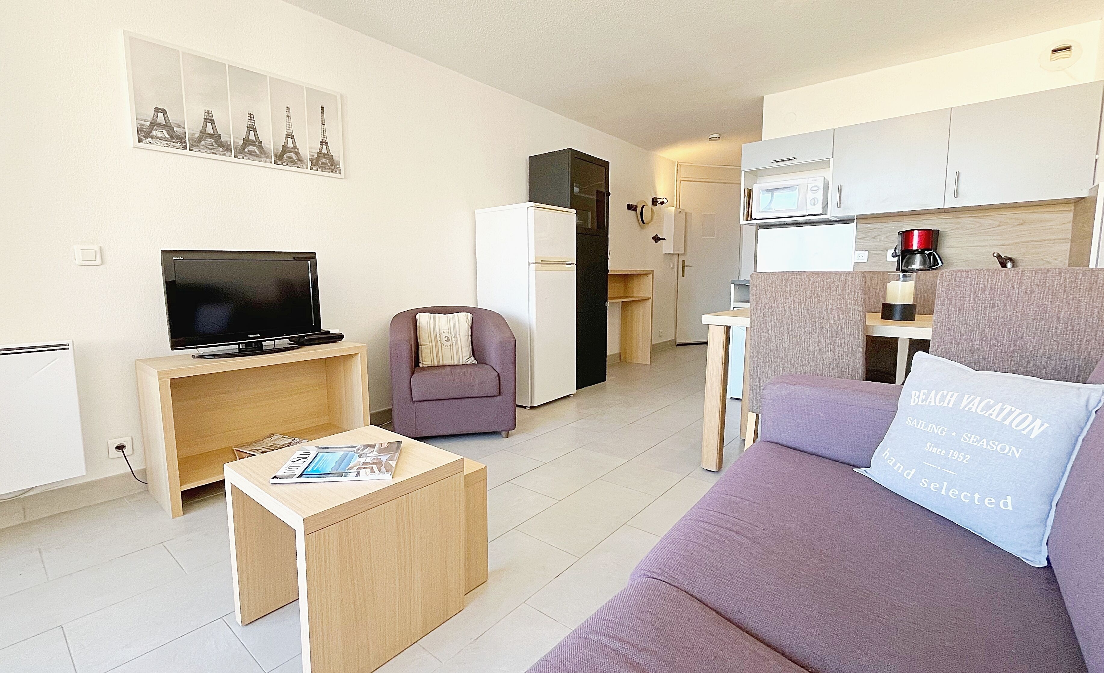 Un espace de vie compact comprenant un téléviseur, un canapé, un petit fauteuil, une table basse et un coin cuisine.