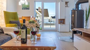 Een moderne woonkamer met een houten tafel, daarop wijnglazen en een fles, een groene bank en zicht op een balkon met een hekwerk en een gebouw op de achtergrond.