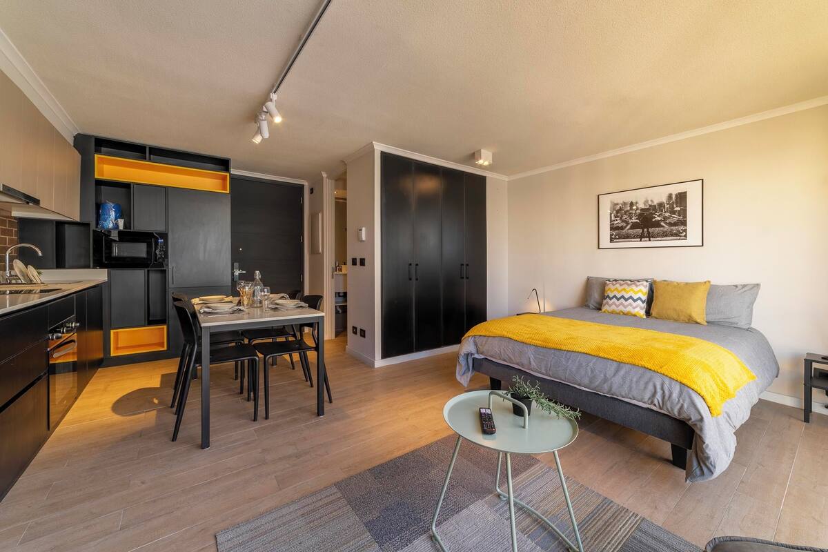 Una habitación de hotel moderna con cama, mesita de noche y una pequeña zona de comedor con mesa y sillas.