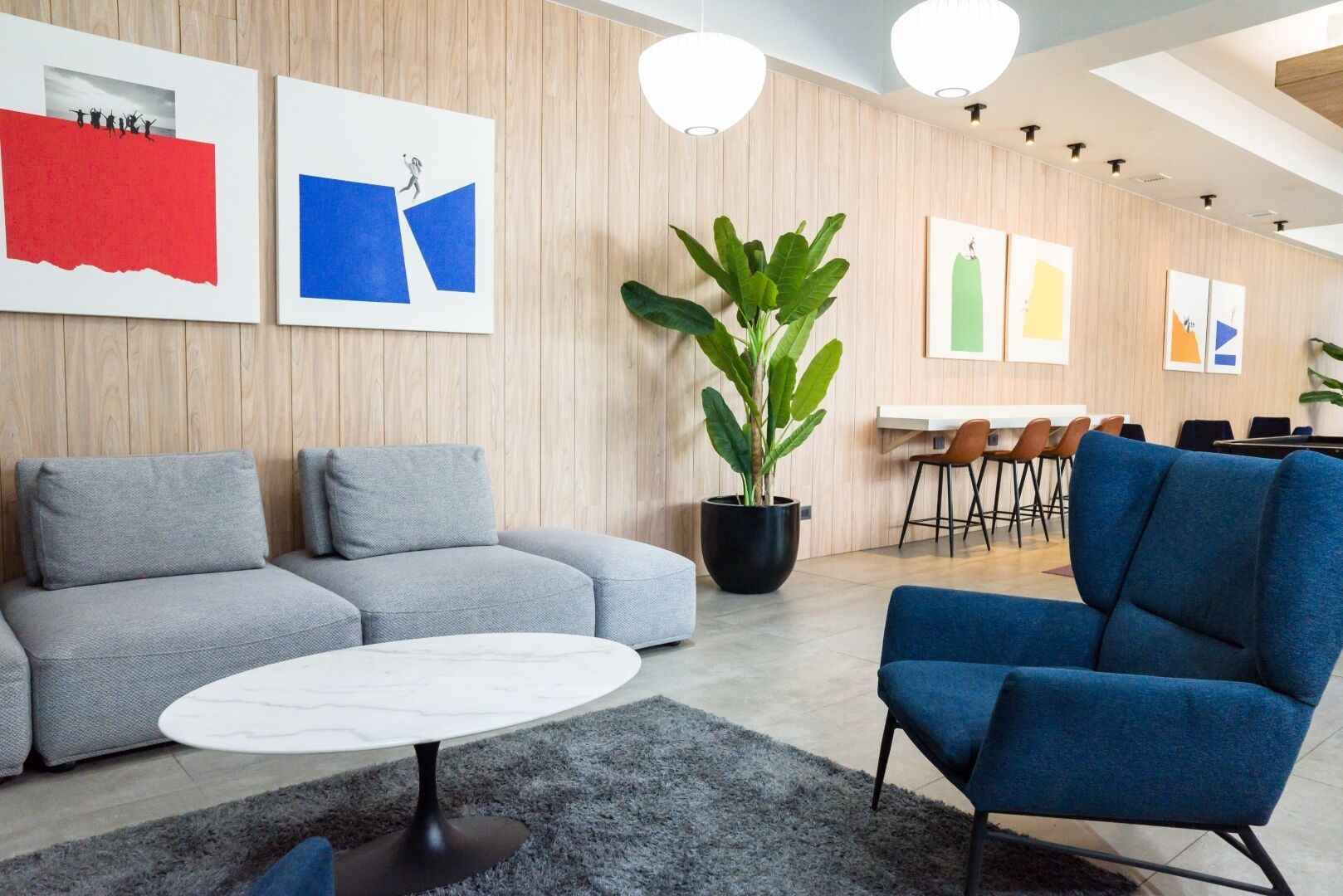 A modern office space with a grey sectional sofa, a blue armchair, a round white table, and a large potted plant.