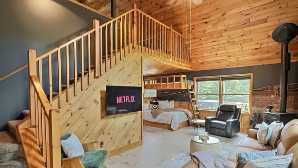 Interior acogedor de una cabaña con una escalera de madera, una cama con un televisor que muestra 'NETFLIX', un sofá y una chimenea.