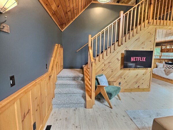 Un escalier en bois, avec une enseigne portant l’inscription « NETFLIX » sur le mur, mène à une pièce où se trouvent un lit et une fenêtre.