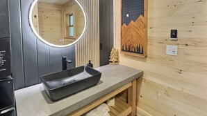 Un baño con pared de madera, un espejo redondo, un lavabo negro y un estante de madera.
