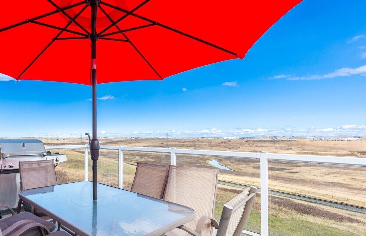 A balcony with a glass table and chairs, a red umbrella, and a view of a railway track and distant landscape.