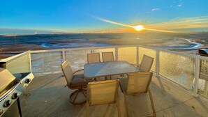 A balcony with a table and chairs, overlooking a beach with the sun setting in the background. - Covington Close (Calgary)