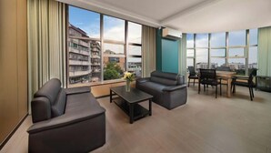 A modern living room with a sofa, a coffee table, and a dining area with chairs and a table. Large windows provide a view of the cityscape. - Anqing Yixin Hotel (Anqing)
