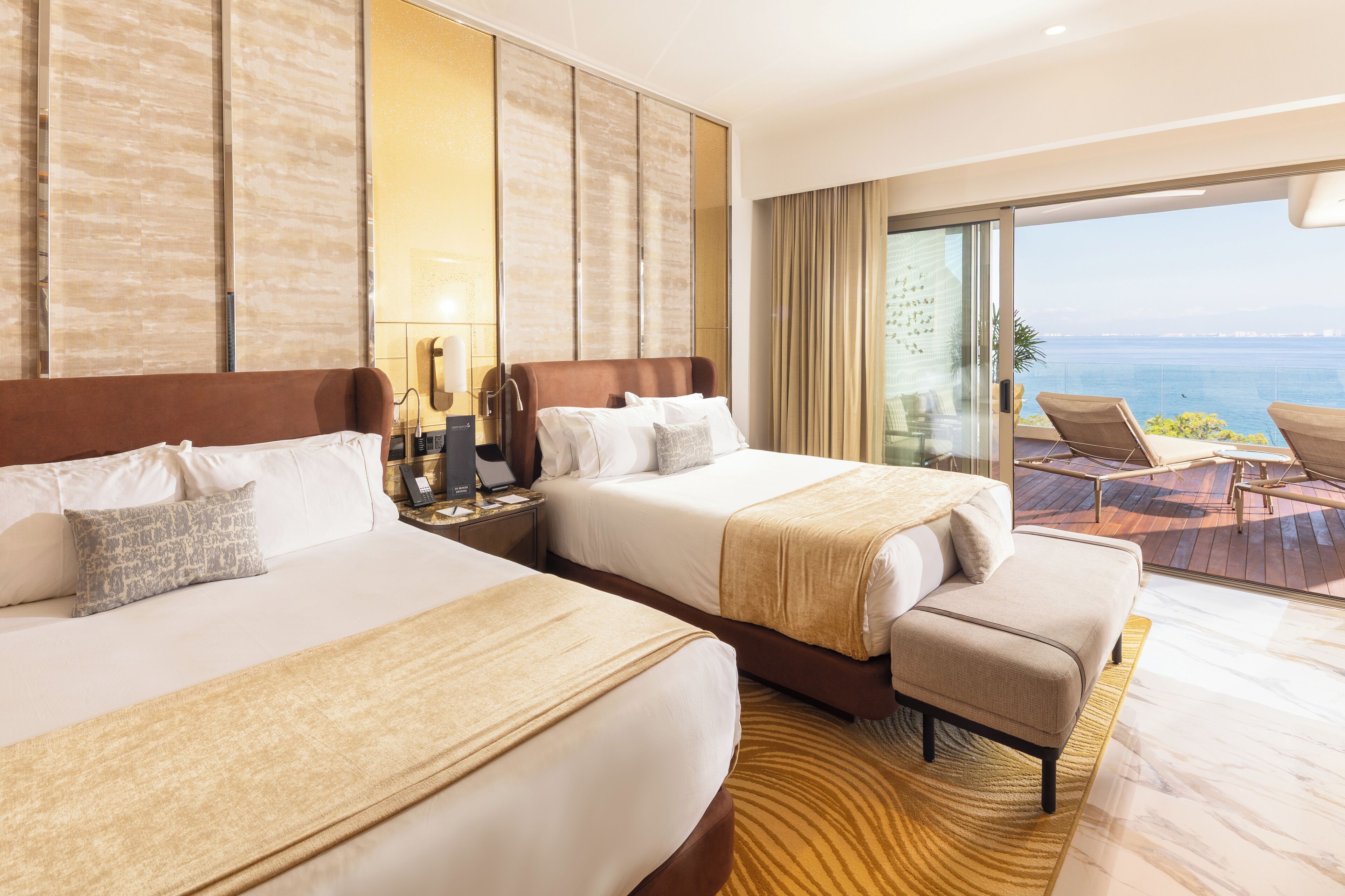 A hotel room with two beds, a sofa, a balcony with a view of the sea, and a large mirror on the wall.