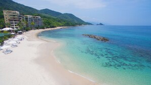 Una playa con agua turquesa cristalina, una orilla arenosa y un hotel de varios pisos con balcones con vistas al mar.
