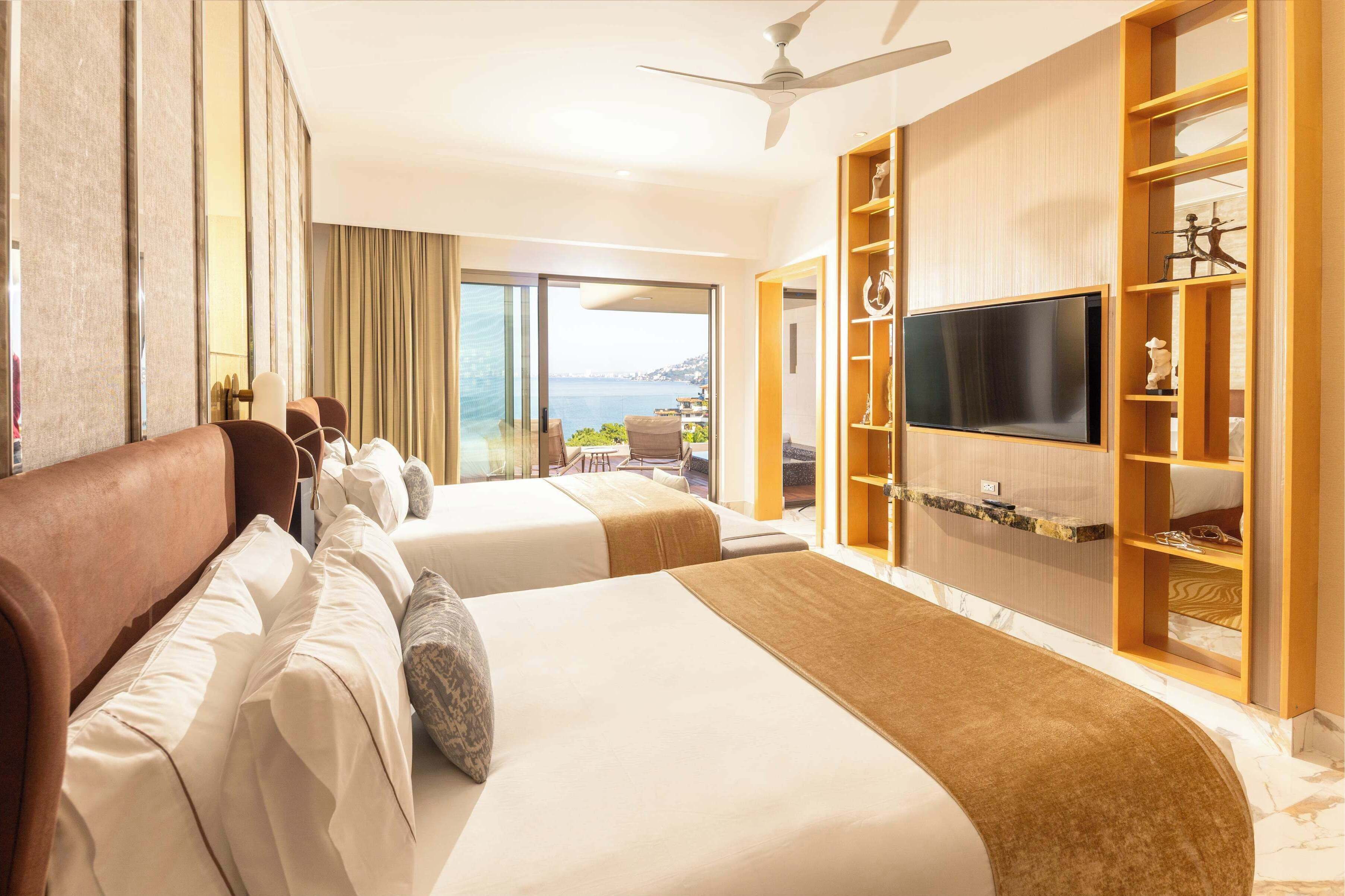 A hotel room with two beds, a TV, a ceiling fan, and a view of the sea.
