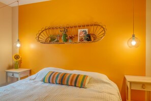 Un dormitorio con cama, mesita de noche con un florero de girasoles y estante de pared con adornos.