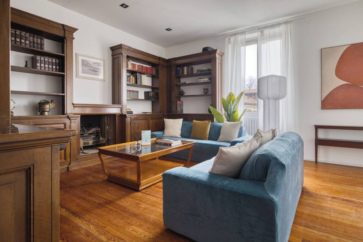 Uma sala de estar com um sofá azul, estantes de madeira, uma mesa de centro e uma janela grande com cortinas.