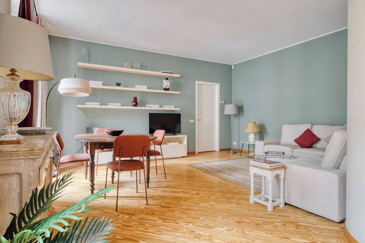 Uma sala de estar com sofá, mesa de centro, mesa de jantar com cadeiras, televisão e estantes com livros e objetos decorativos.