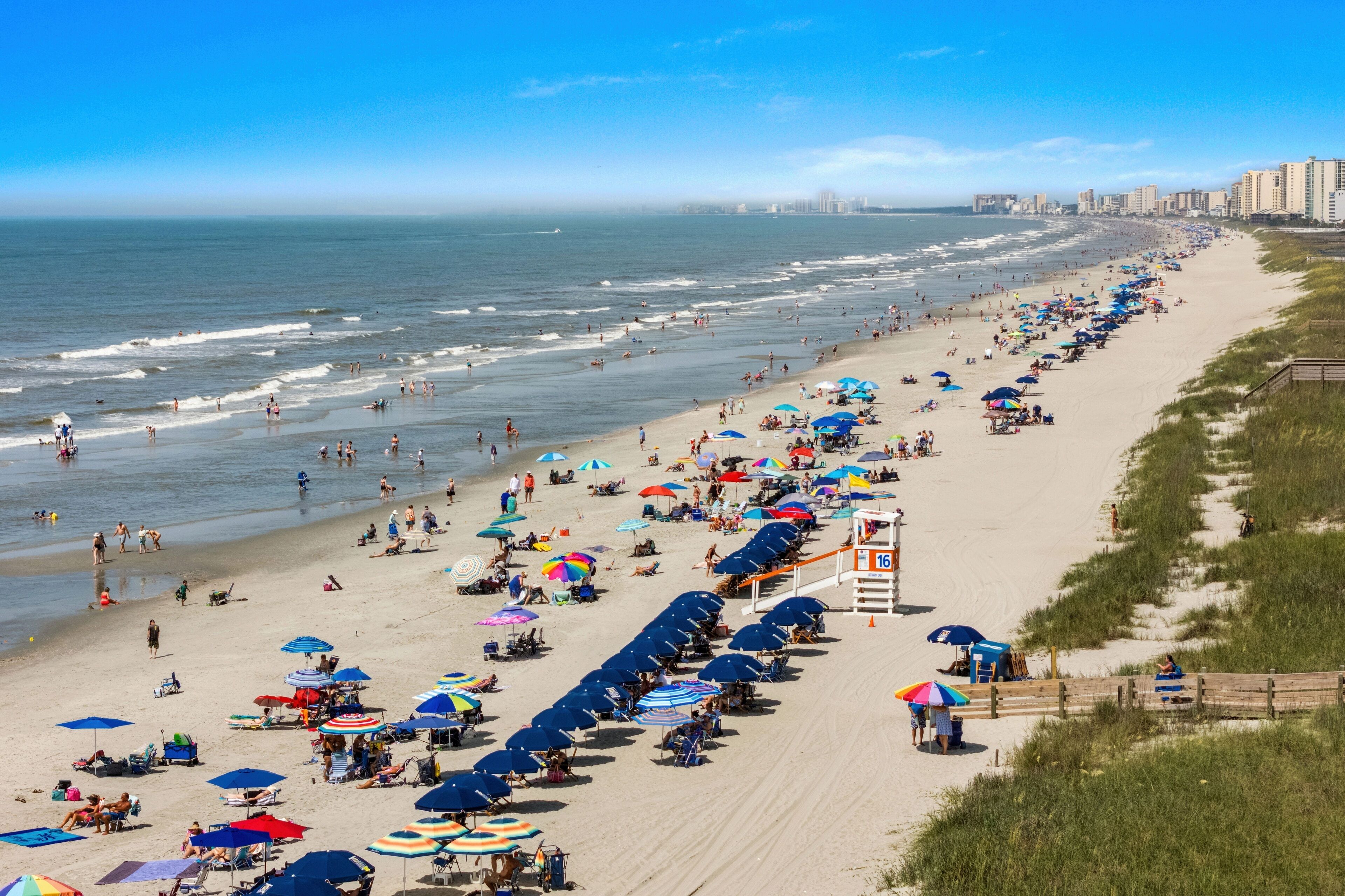 A beach with rows of umbrellas and beach chairs, a lifeguard tower, and a boardwalk along the shore.