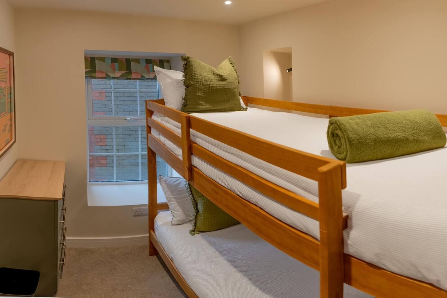 A bunk bed with a green towel, a window with a view of a brick wall, and a small desk with drawers.