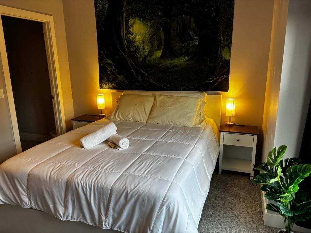 Een netjes opgemaakt bed met een witte dekbedovertrek en kussens, twee nachtlampjes, een nachtkastje en een groot wandtapijt met een bosmotief.