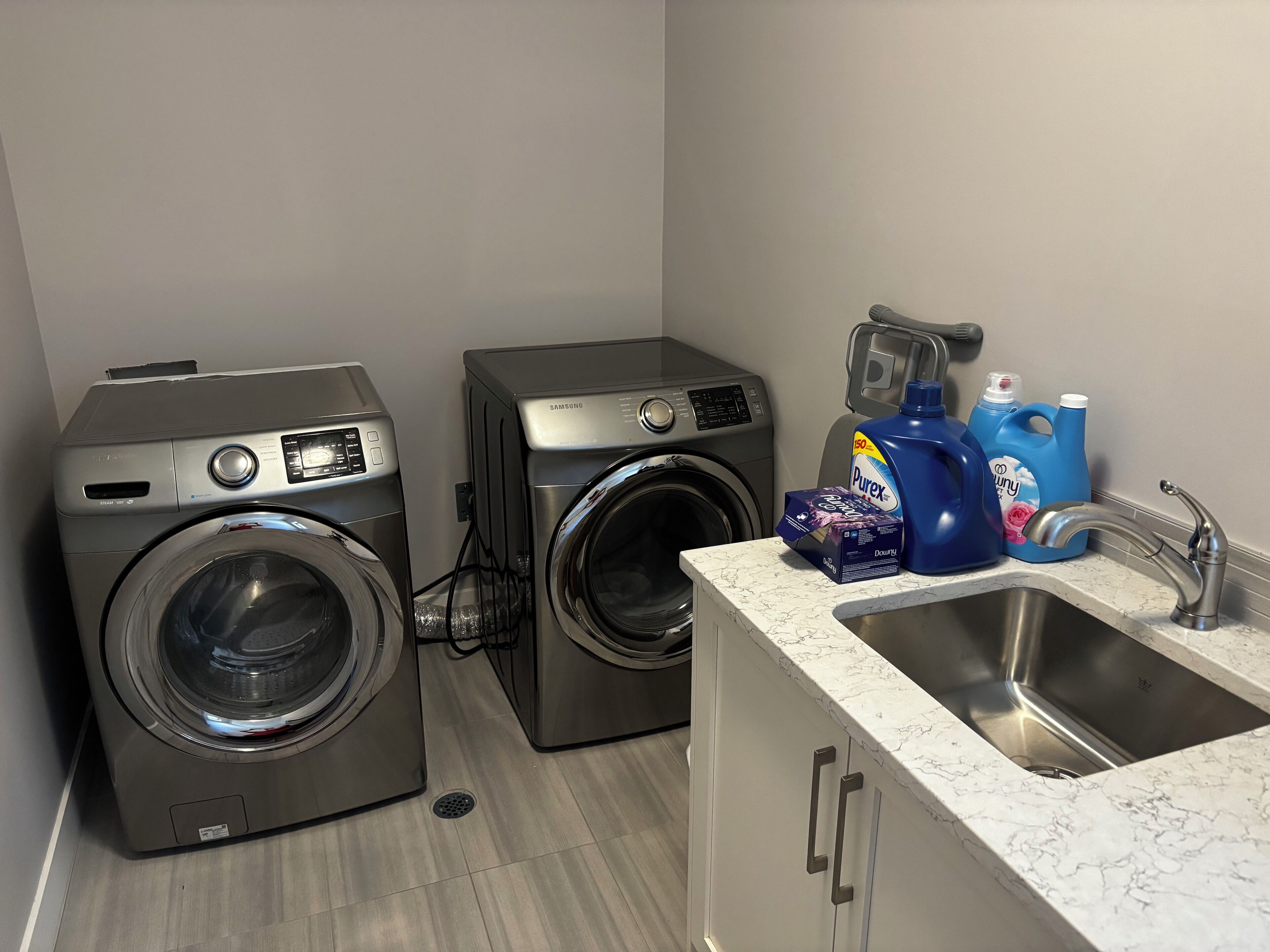 A laundry room with two front-loading washing machines, a sink with a marble countertop, and various cleaning supplies.
