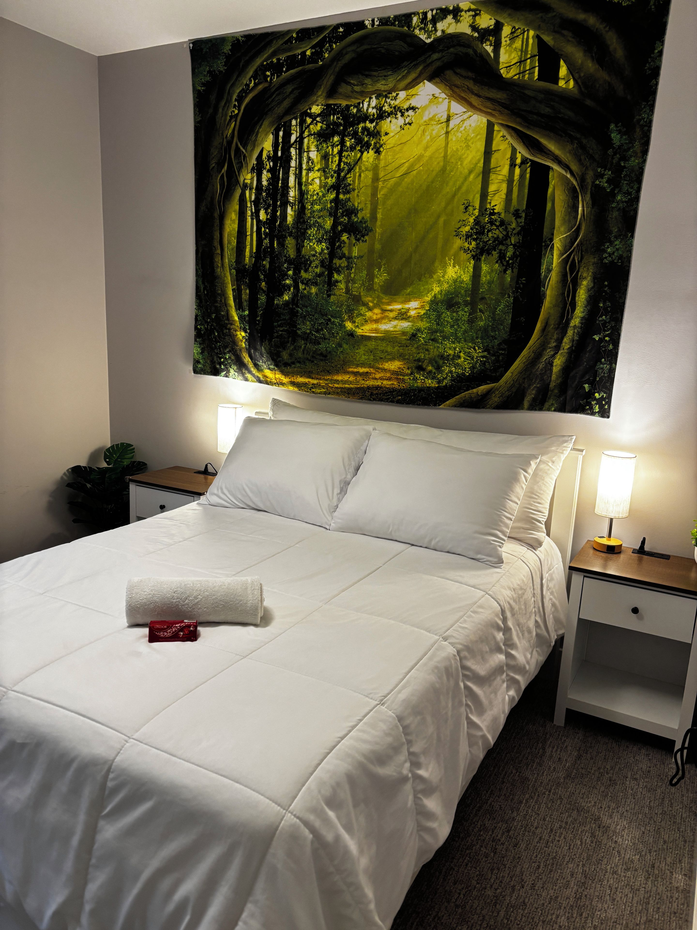 Una cama bien hecha con ropa blanca, un mural temático de bosque y lámparas de noche.