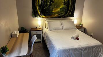 A hotel room with a bed, bedside tables, a desk, a chair, and a wall-mounted painting of a forest scene.