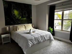 A bedroom with a large bed, a nightstand, a window with curtains, and a wall tapestry.