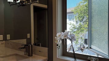 A bathroom with a marble countertop, a sink, a mirror, a window with a view of greenery, and a potted orchid.