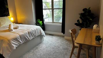 Una cama bien hecha con una almohada y una toalla enrollada, una mesita de noche con una lámpara, una ventana con vistas a edificios, y una pequeña mesa con una planta.
