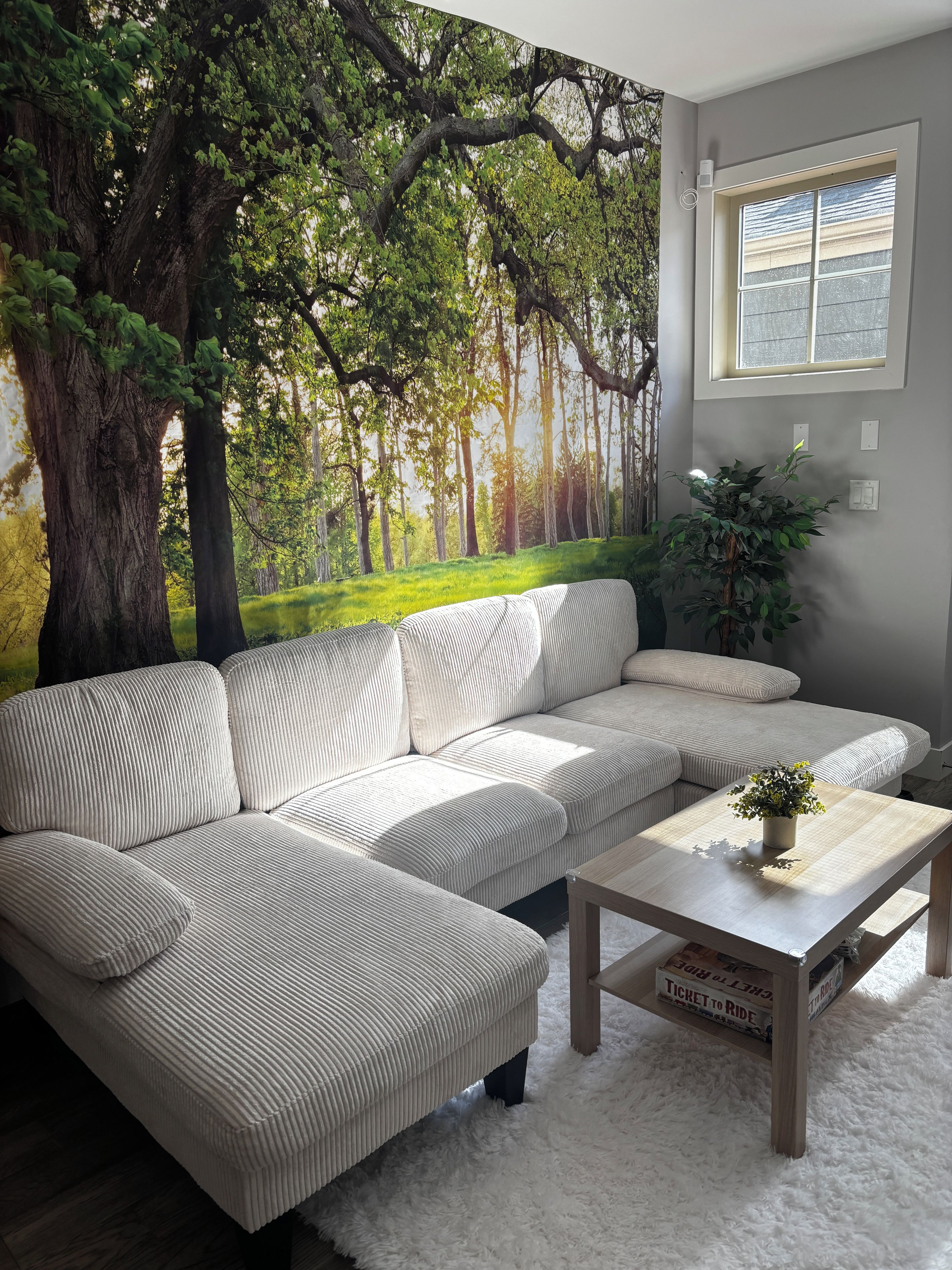 Sala de estar com sofá seccional, mesa de centro e um grande mural na parede retratando uma floresta.