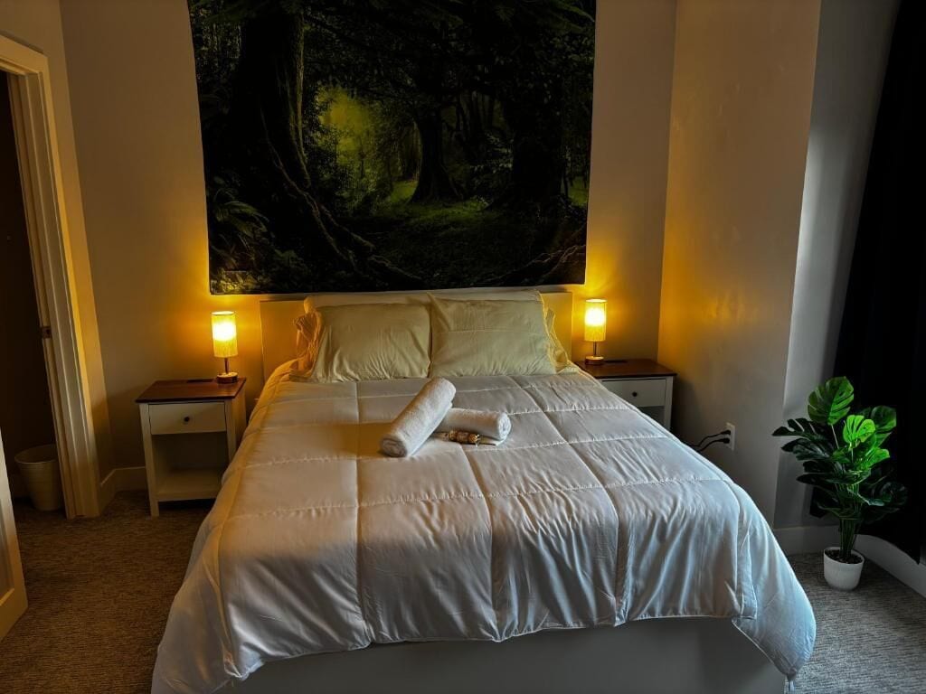 A neatly made bed with a white duvet, two bedside lamps, a large forest-themed tapestry on the wall, and a potted plant on the floor.