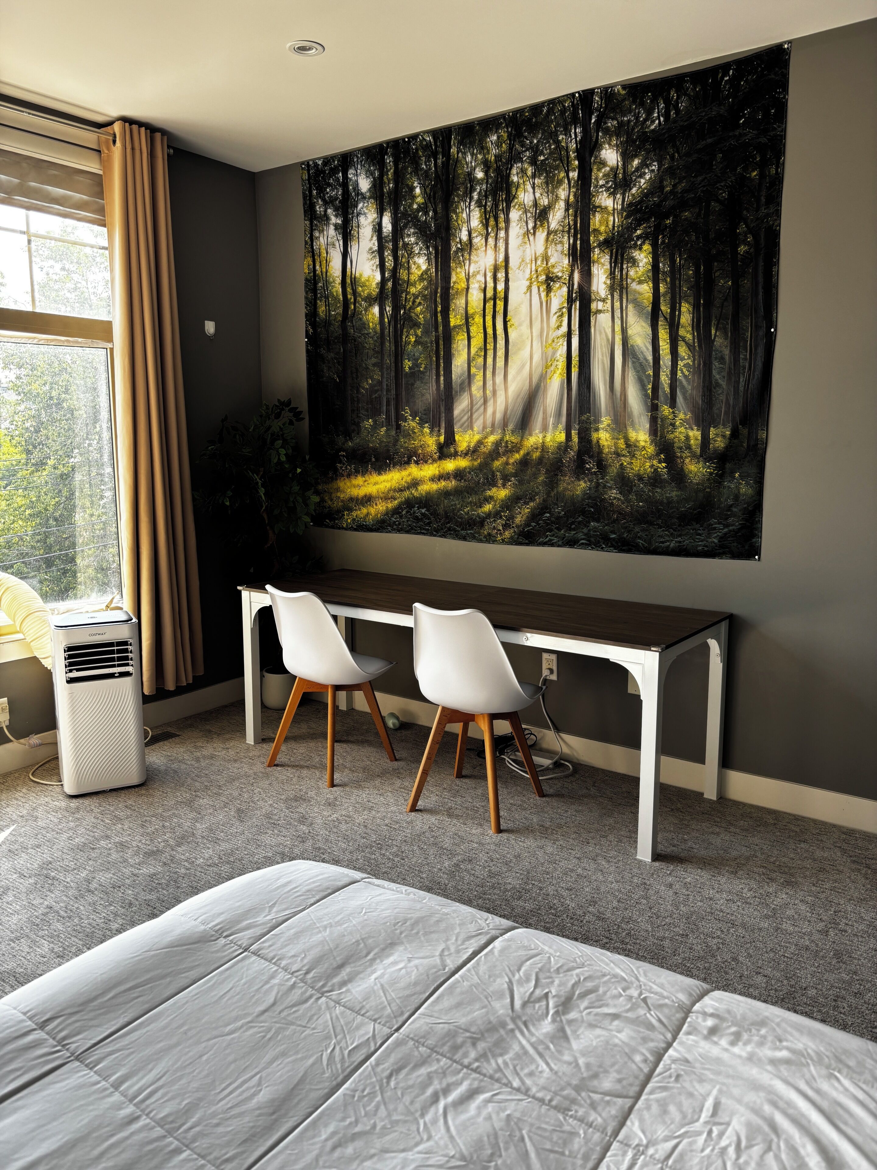 Un dormitorio con una cama, un escritorio y dos sillas, un mural grande con motivos forestales y una ventana con cortinas.
