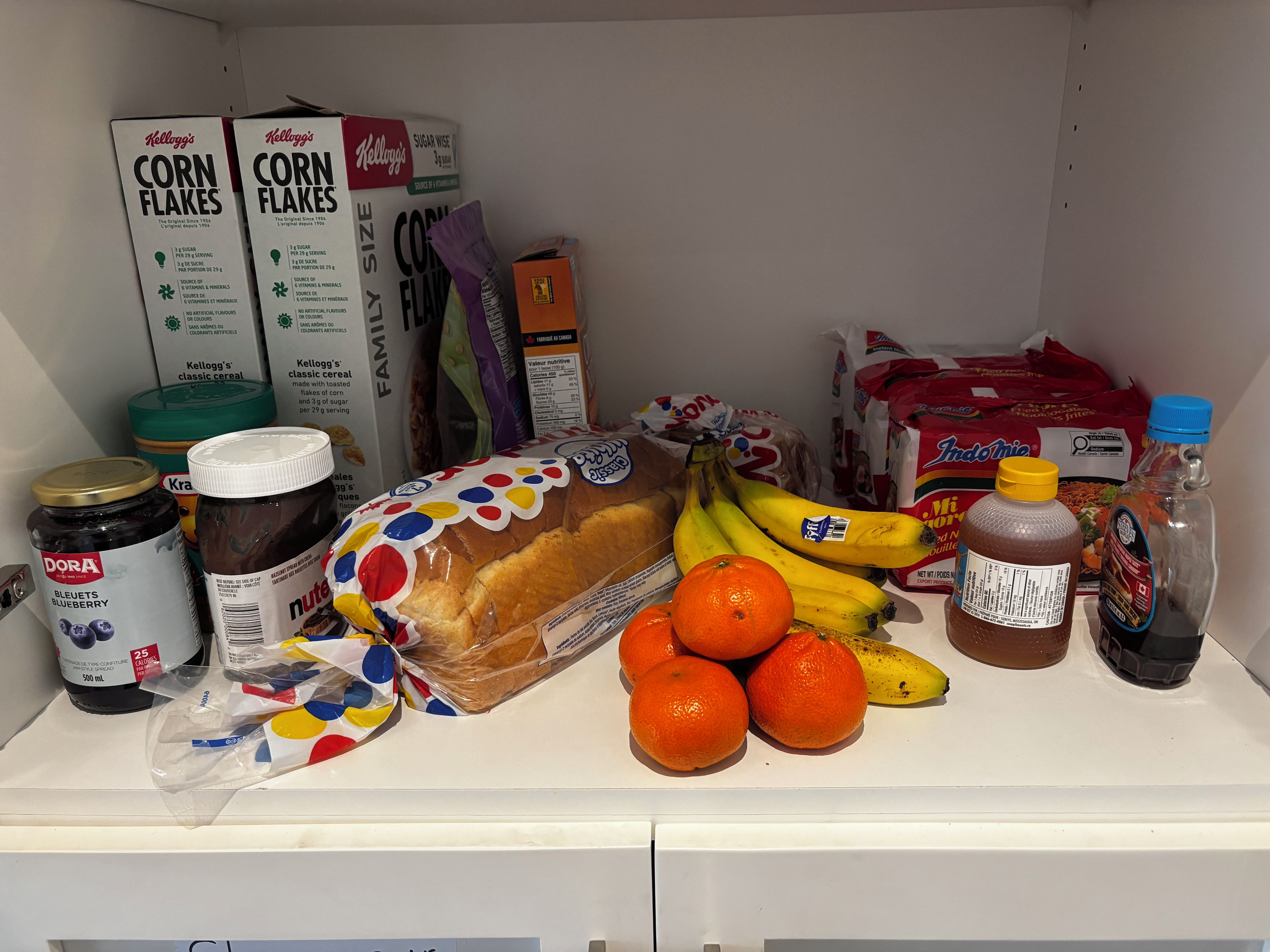 A pantry with various food items including Kellogg's Corn Flakes, bread, bananas, oranges, and jars of preserves.