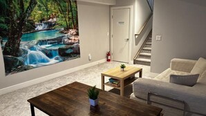 A living room with a large wall mural, a staircase, a sofa, a coffee table, and a small table with a plant. - Nature Home near Downtown and 17th Av (Calgary)