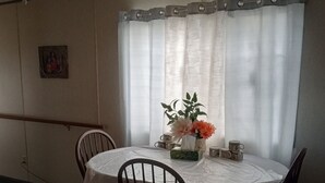 A dining area with a round table covered by a white tablecloth, surrounded by four wooden chairs. There is a vase with flowers on the table, and a window with sheer curtains behind it.
