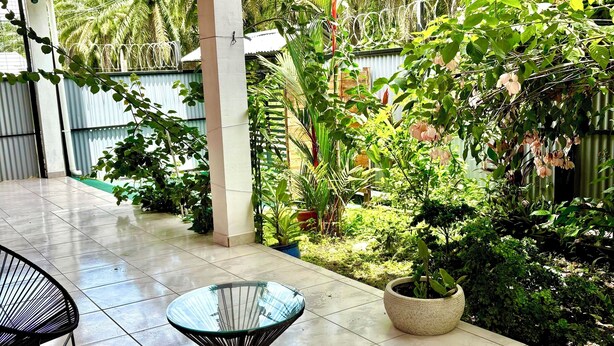 Un patio cubierto con suelo de baldosas, dos sillas de metal negro, una mesa de vidrio y una variedad de macetas con plantas y follaje.