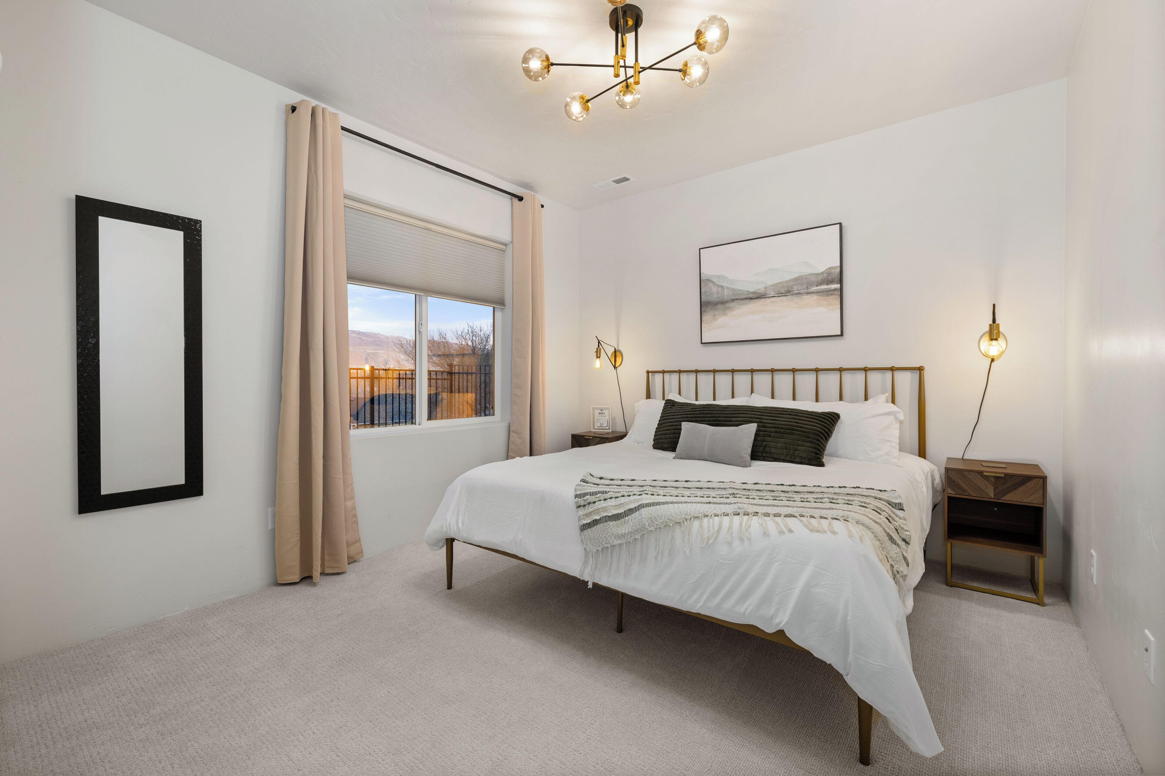 A bedroom with a large bed, a window with curtains, a bedside table, and a wall-mounted light fixture.
