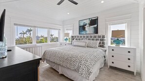 Ein Schlafzimmer mit einem Bett, einem Deckenventilator, einem Fernseher, einem Fenster mit Blick auf Palmen und einem Nachttisch mit Schubladen.