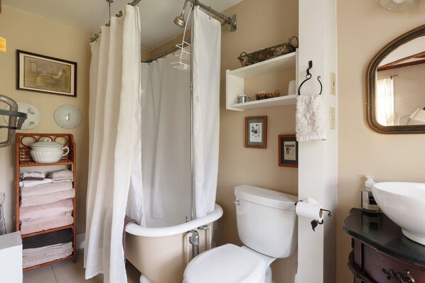 Ein Badezimmer mit Toilette, Waschbecken und Duschvorhang. Es gibt Regale mit Handtüchern und ein gerahmtes Bild an der Wand.
