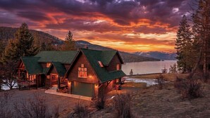 A cabin with a green roof, wooden exterior, and a dock extending into a lake, surrounded by mountains and trees. - Bottle Bay View Lodge – 21-Acre Lakefront Retreat (Sagle)