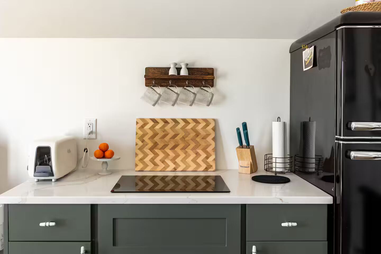 Una cocina con encimera blanca, refrigerador negro, tabla de cortar de madera, bloque para cuchillos y tostadora.