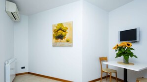 Une pièce avec un radiateur blanc, un tableau représentant des arbres, un téléviseur et un vase rempli de tournesols.