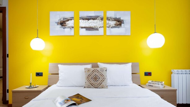 Une chambre d’hôtel avec un mur jaune vif, une tête de lit en bois, du linge de lit blanc et deux tables de chevet avec des lampes.