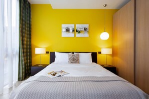 Une chambre d’hôtel avec un mur jaune vif, un lit avec une literie blanche, deux lampes de chevet et un luminaire suspendu.